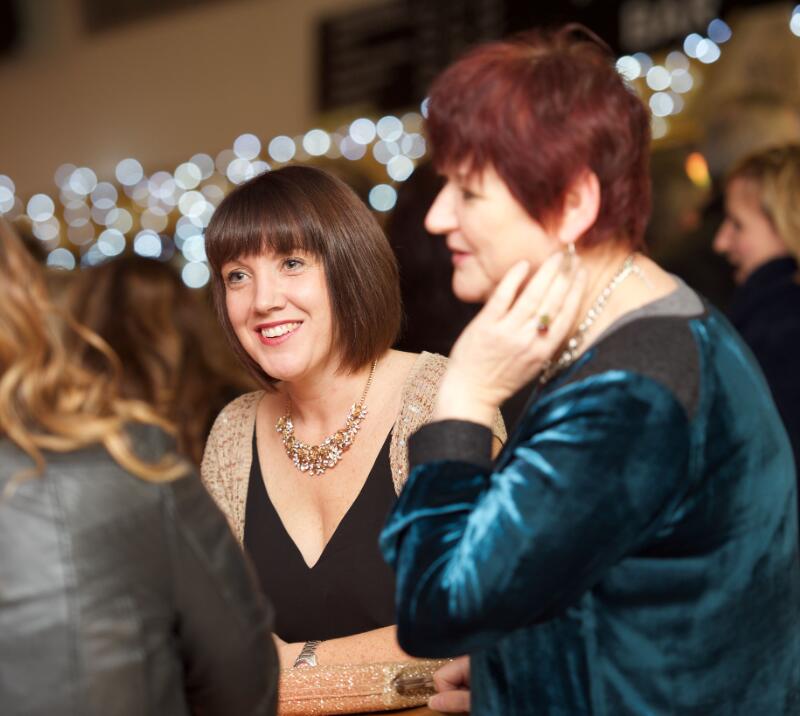 Festively dressed women chat in front of sparkly lights.